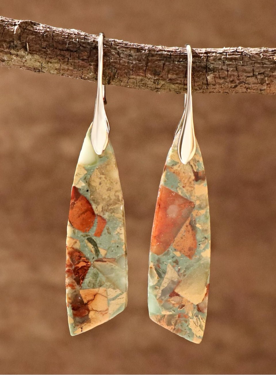 Pair of earrings with a natural, multicolored design hanging on a branch against a brown background.  (Silver)