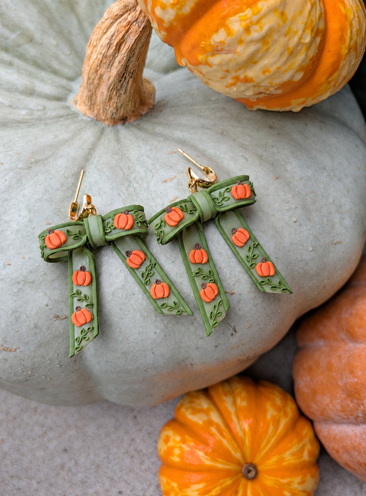 Pair of sage-green bow dangle earrings with six orange pumpkins on each bow, green vines, and gold-toned hardware