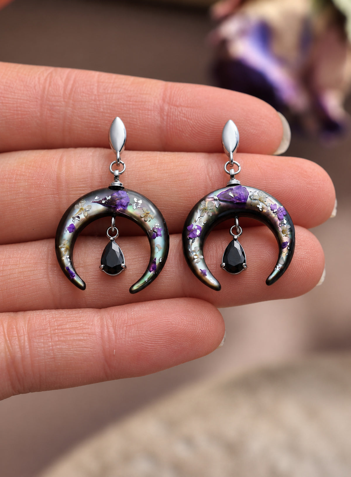 Pair of silver-finish earrings with purple flowers on dark crescents and hanging black teardrop stones.